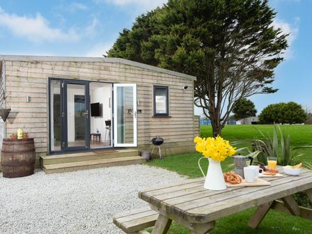 A wooden cabin with open glass doors and a picnic table with flowers and breakfast items at Bushwood Lodge in Wendron near Helston