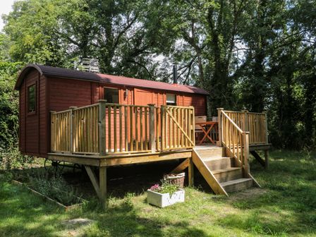 A wooden shepherds hut with a raised deck and stairs in a grassy area surrounded by trees at The Foxes Den Shepherds Hut in Oldbury-On-Severn