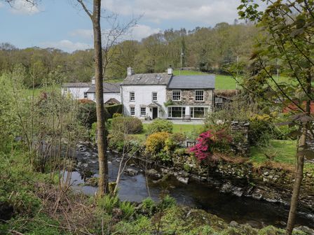 A stone and white house beside a river with trees and shrubs at Beck Cottage in Satterthwaite