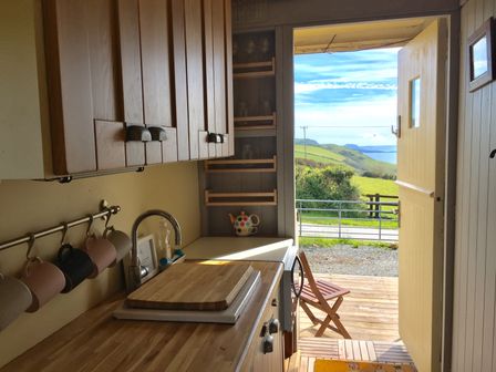 A kitchen with wooden cabinets a sink mugs hanging on a rail and a door open to a deck with a chair overlooking green hills and water at Sea the Stars in New Quay