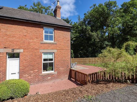 An exterior view of a brick house with two windows and a white door surrounded by a garden and wooden fence at 2 Twizel Station Cottage Cornhill-on-Tweed near Norham