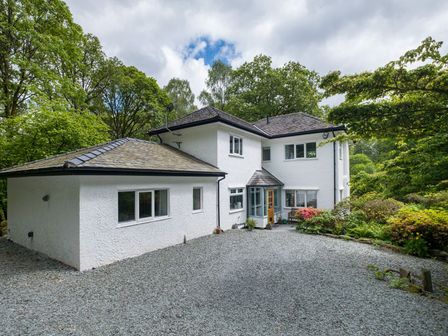 A white two-story house with a gravel driveway surrounded by trees and shrubs at Bridge Howe in Ambleside