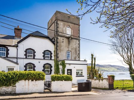 A stone tower attached to a white house by a waterfront with bushes along the wall at The Old Coastguard Tower Buncrana County Donegal