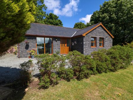 A stone house with wooden doors and windows surrounded by green bushes and trees at Ty Gareg in Llanddaniel near Gaerwen