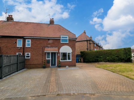 A semi detached brick house with a tiled roof and paved driveway with hedges and grass at Corner Cottage in Bridlington