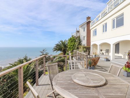 A wooden outdoor dining table with chairs on a balcony overlooking the ocean with a white building and palm trees at Seahaze in Downderry