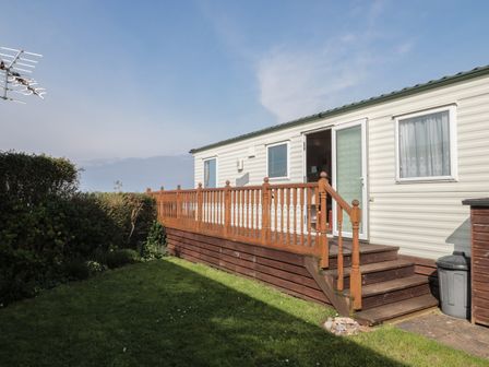 An exterior view of a mobile home with a wooden deck and steps leading to a door with green grass and bushes nearby at Bayview 23 in West Quantoxhead