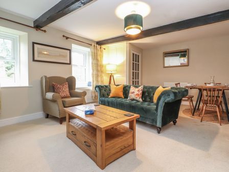 A living room with a green sofa yellow cushions a wooden coffee table an armchair two windows and a dining table at Ash Tree Cottage in Hayton near Brampton Cumbria