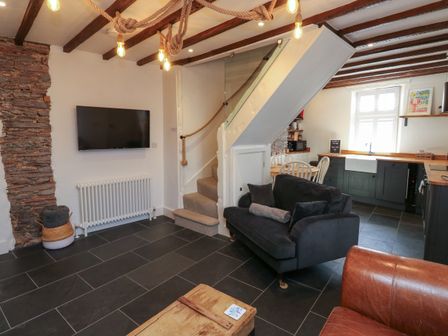 A living area with a wall-mounted TV a staircase a black sofa a dining table and a kitchen with a window at The Nook in Brixham