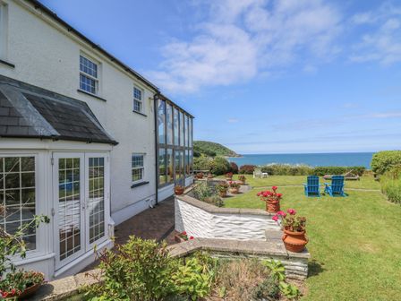 A white house with a glass enclosed area and flower pots on a terrace overlooking a lawn with blue chairs and ocean view at Pen y Graig in Aberporth