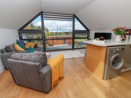 A living area with two sofas near large triangular windows and a kitchen island with a washing machine at Cuckoo's Rest in Penryn