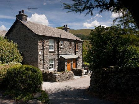 A stone cottage with two wooden doors surrounded by greenery and a stone wall at The Cosy Peacock in Troutbeck