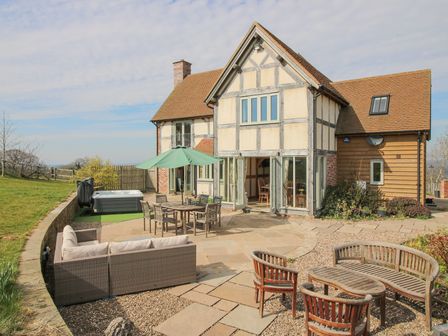An outdoor patio area with seating furniture including a sofa set, dining table with umbrella, wooden chairs and table next to a house at Cuckoo Pen Cottage Eastham near Tenbury Wells