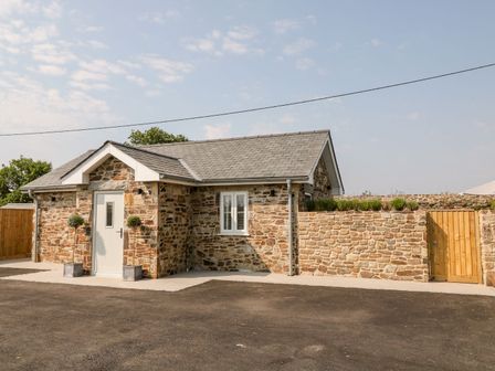 A stone cottage with a white door and window next to a stone wall and wooden gate at Pear Tree Cottage in St Mellion near St Dominick