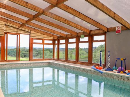 An indoor swimming pool with wooden beams and large windows overlooking trees with pool toys on the side at Skiber Long in Bray Shop near Launceston