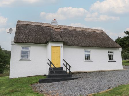 A white cottage with a thatched roof yellow door and black steps surrounded by grass and gravel at Bofin in Oughterard County Galway