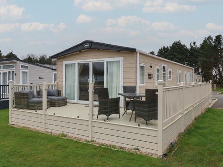 A beige static caravan with a fenced wooden deck containing outdoor seating and a table at Whispering Willows in Cayton Bay