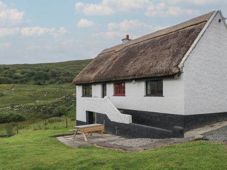 A white stone cottage with a thatched roof and outdoor bench near green hills at Corrib Near Oughterard County Galway