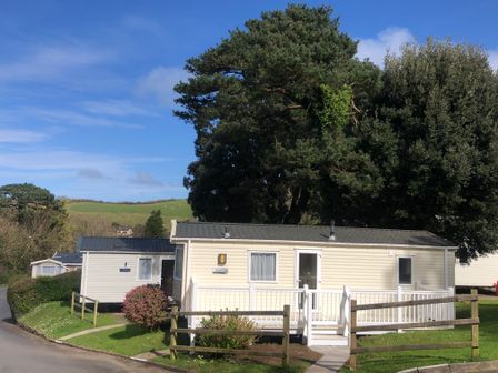 A cream mobile home with white railings surrounded by trees and greenery at Ocean View in Combe Martin