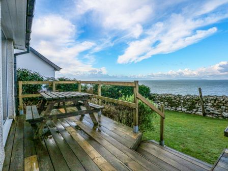 A wooden deck with a picnic table and view of a stone wall grass and ocean at Ael Y Don in Llandanwg