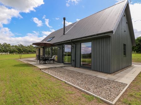A house with a gray metal roof and dark vertical siding with an outdoor table umbrella and chairs on a patio at Strawberry Cottage in Wester Essendy near Dunkeld