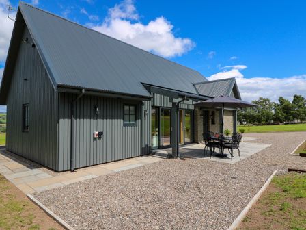 An outdoor patio area with a table, chairs, and umbrella beside a modern building with grey cladding and metal roof at Bramble Cottage in Wester Essendy near Dunkeld