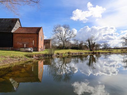 A pond in front of brick and black barns with leafless trees and clouds reflected at Brook Farm in St Nicholas near Halesworth