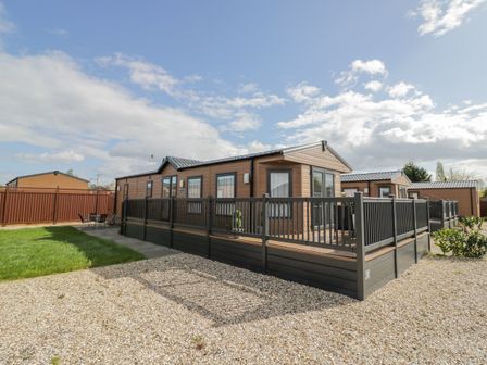 A brown static caravan with a black deck fence on a gravel and grass plot at Long Acres Lodge in Evesham