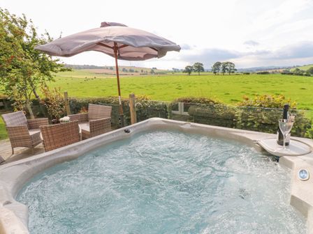 An outdoor hot tub with bubbling water next to a patio table with four chairs and a large umbrella overlooking a green field at Plot 32 High Hesket near Armathwaite