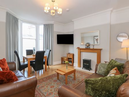 A living room with brown leather sofas green and orange cushions a wooden coffee table a fireplace a dining table with black chairs and large windows at Baystones in Keswick