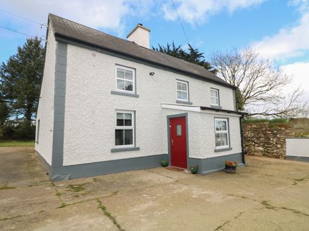 A two-story house with white walls gray trim and a red door surrounded by a paved area at Curragh Cottage in Rathimney near New Ross County Wexford