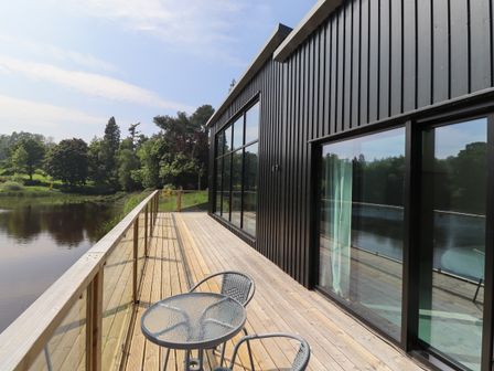 A wooden deck with glass railing and a metal table with two chairs outside a black wooden building overlooking a body of water at Otters Cove in Belford