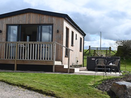 An exterior view of a wooden cabin with a small porch and railing a hot tub with two chairs next to it on a patio at Cae Pren in New Mills near Llanfair Caereinion