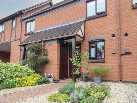 The front entrance of a brick house with a covered porch and various plants and shrubs around the pathway at Wren Cottage - Heritage Mews in Stratford-Upon-Avon