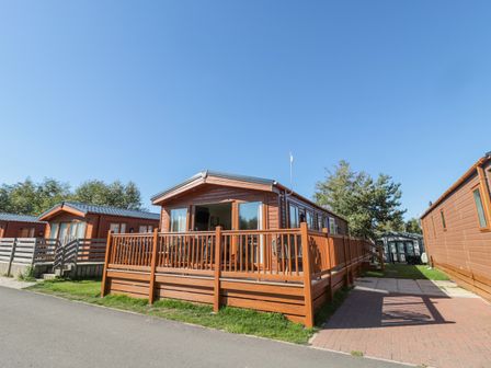 A wooden holiday lodge with a deck and railings on a paved path at Acorn Lodge in Felmoor Holiday Park near Felton