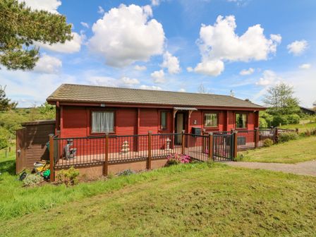 A red wooden lodge with a fenced porch and a surrounding grassy area at Moonlake Lodge in Woolsery near Woolfardisworthy