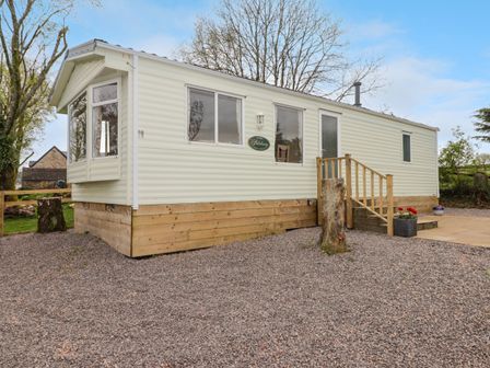 A white mobile home with wooden skirting and small stairs on a gravel lot with trees in the background at Grandads Shack in Trevethin near Pontynewynydd