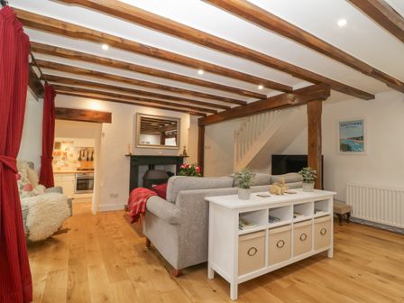 A living room with wooden beams on the ceiling a gray sofa a white shelving unit with plants and a decorative rabbit and an adjacent kitchen visible through an open doorway at 4 Barton Cottages Down Thomas near Plymouth