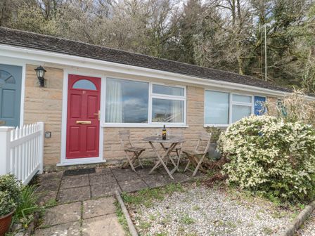 A small patio area with a wooden table and three chairs outside a house with a red door and large windows at 27 Fernhill Heights in Charmouth