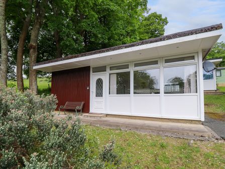 The exterior of a small building with large windows and a white door next to a bench and bushes at Hill View in Bryncrug near Tywyn