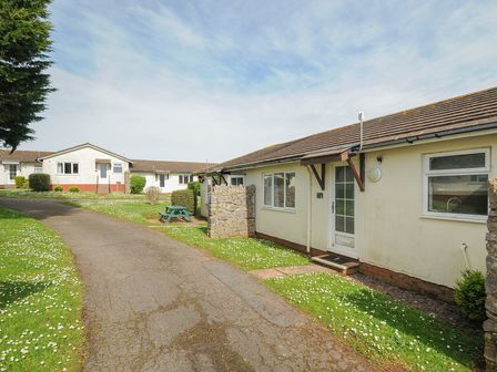 A pathway running between single-story bungalows with green lawns and a picnic table at Chalet 27 at Landscove Holiday Park in Brixham