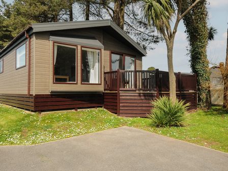 An exterior view of a wooden lodge with large windows and a small deck surrounded by grass and trees at Lodge 15 at Landscove Holiday Park in Brixham
