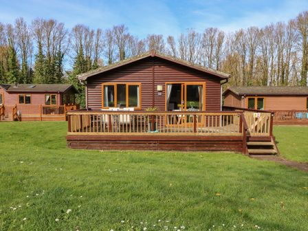 A wooden cabin with a deck and outdoor furniture surrounded by grass and trees at 3 The Glade in St Minver Rock