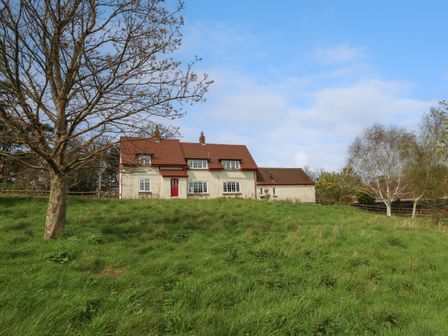 A house with red roof and red door behind grassy hill with trees at Rempstone Farmhouse in Corfe Castle
