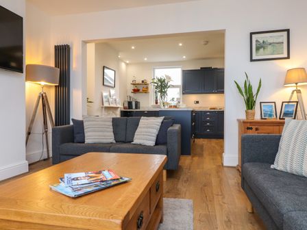 A living room with two gray sofas a wooden coffee table and lamps leading into a kitchen with dark cabinets at Dolfor in Cemaes Bay
