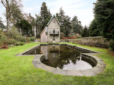 A small stone house with a green roof and a clock above the door beside a stone-lined pond surrounded by grass and trees at The Folly in Belford
