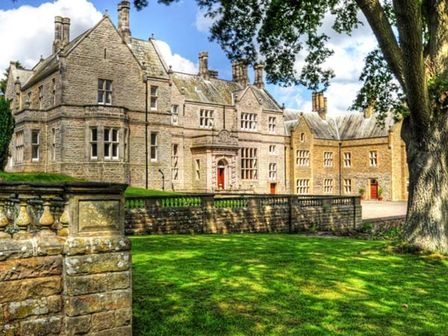 A large stone mansion with multiple chimneys and windows surrounded by a stone fence and grassy lawn at The Hall in Belford