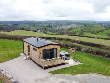 A small wooden cabin with a patio and hot tub surrounded by fields with sheep at Cae Bedw in Llanfair Caereinion