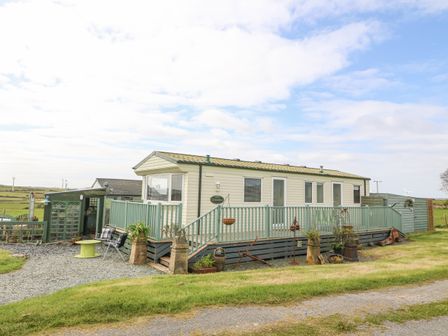 A caravan with a wooden deck and outdoor seating surrounded by grass at The Caravan @ Lletty'r Wennol in Cemaes Bay