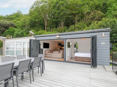 An outdoor dining table and chairs on a deck with an open living area and bedroom at 100 Hillside Villas in Looe